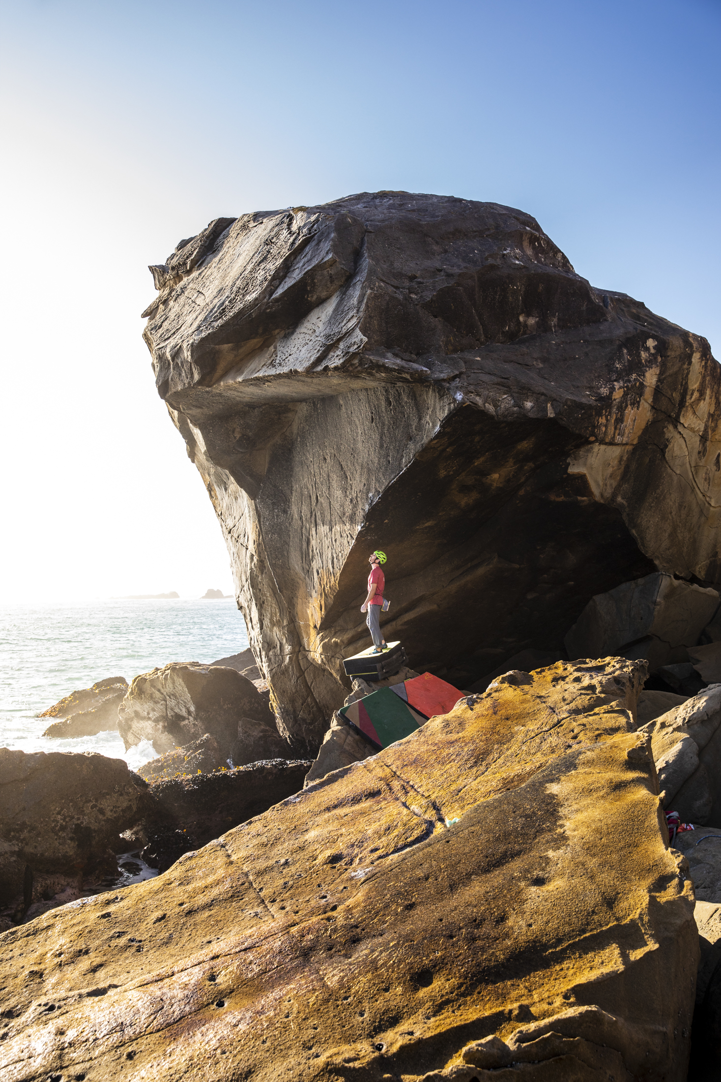 Photograph at sunset of Kevin Jorgeson standing on a stack of pads at the base of an unclimbed highball bouldering problem in the surf of California's Sonoma Coast.